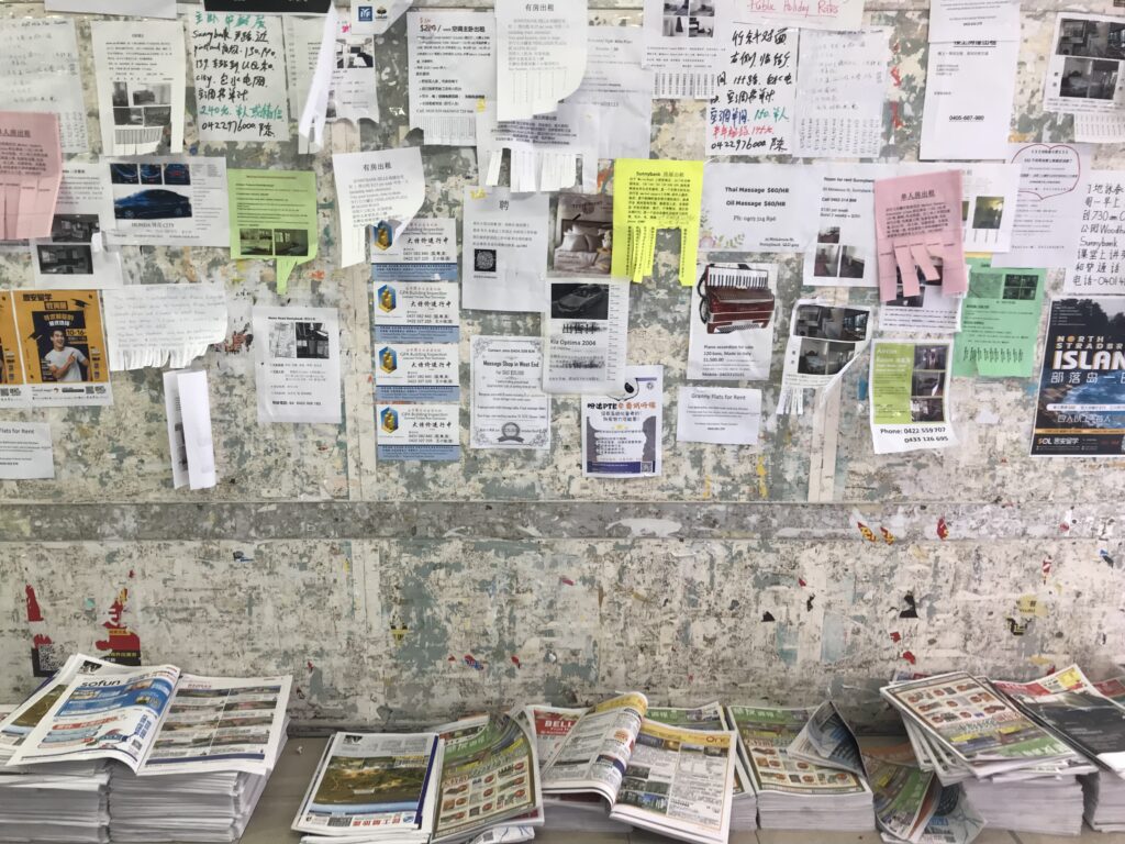 Sunnybank公告墙正面，三分之二贴满浅色传单，墙根报纸堆