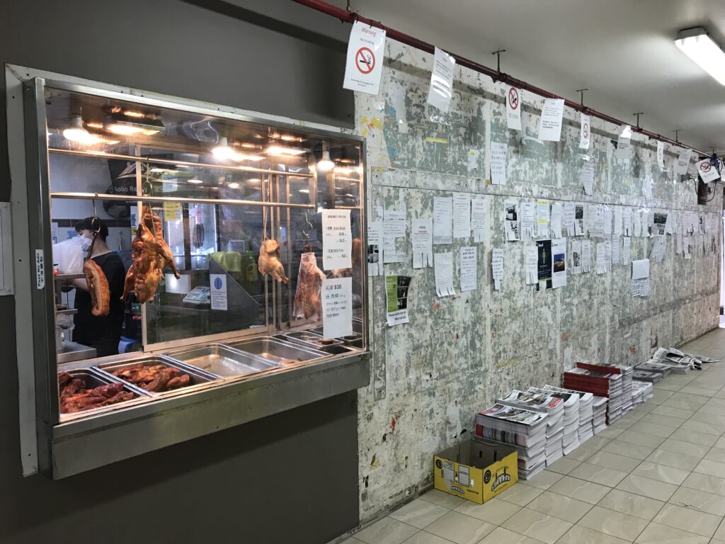 Sunnybank墙烧腊店一端，两排传单，墙根报纸堆，烧腊橱窗挂几只鸭