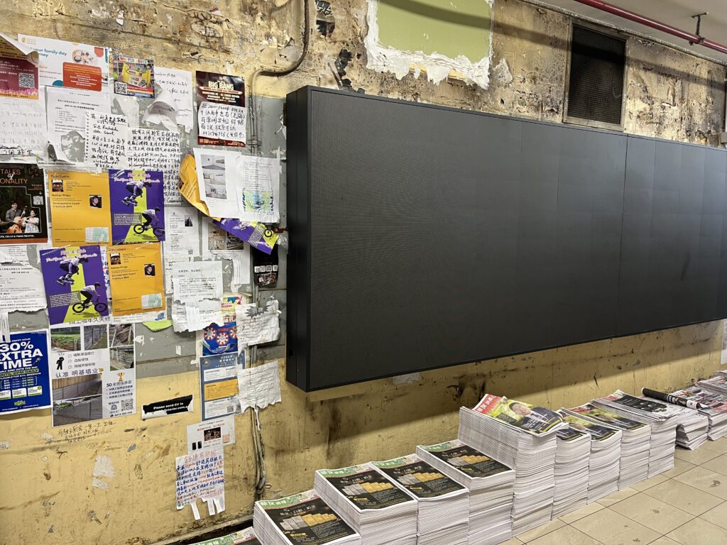 Sunnybank墙裸砖段，黑色LED屏占大半画面，墙根报纸堆，顶部管线交错