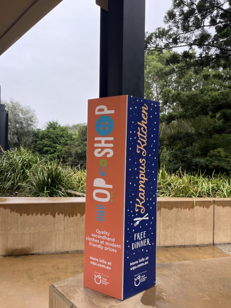 UQ湖边公交站通往校园的步道旁，另一块学生会彩色标识牌，宣传二手衣物平价商店和免费晚餐活动。