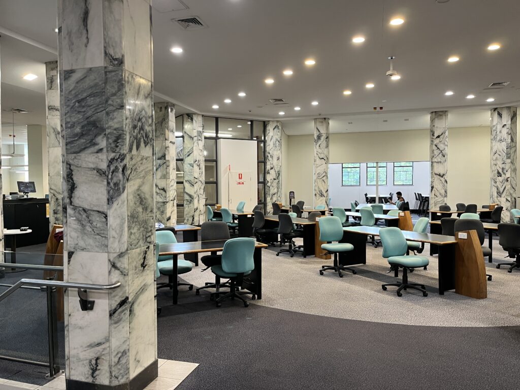 从昆士兰大学中央图书馆Great Court入口的玻璃门内拍摄，可见主咨询台和供学生使用的学习桌。图书馆大厅宽敞明亮，是学生常用的学习空间。