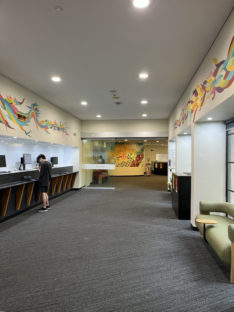 昆士兰大学图书馆下层走廊，通往玻璃门后的电脑区和数据库查询区。走廊左侧有快速查询电脑，右侧落地窗配沙发休息区。凤凰壁画从楼梯延伸至沿墙上端，对面有其他彩色壁画。