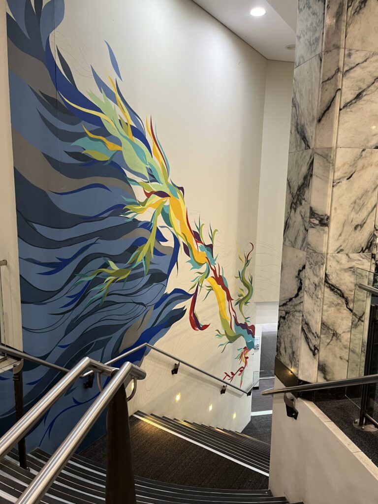 昆士兰大学中央图书馆楼梯，向下通往底层大厅。白色楼梯围绕黑白大理石柱旋转，不锈钢扶手靠白墙。墙上有抽象彩虹色壁画，蓝尾舒展，在中国文化视角下形似凤凰。作品名为"知识重生"。