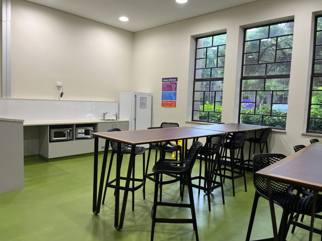 昆士兰大学中央图书馆用餐区，设有微波炉、水槽、冰箱、两排桌椅和回收垃圾桶。墙上有社区公告板，三扇落地窗引入自然光，窗外可见绿色灌木和对面建筑。