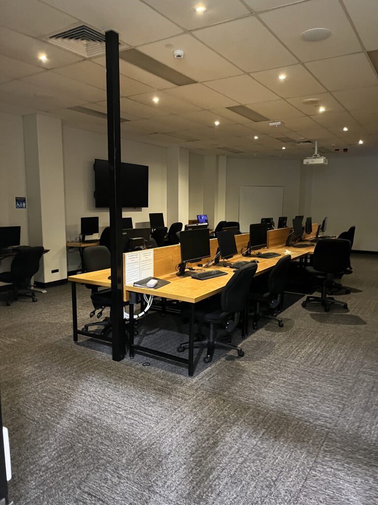 昆士兰大学中央图书馆电脑培训室，暖黄色灯光，墙边排列书桌和台式电脑，中央有两排电脑桌。前方有投影仪和壁挂大电视屏幕。