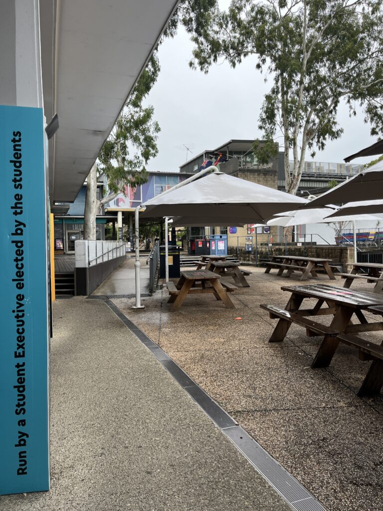 从通往UQ湖边公交站的路回望昆士兰大学学生会综合楼，可见底层食堂、户外用餐区、奶茶店。右侧楼下是Red Room学生酒吧。