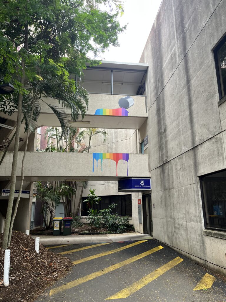 昆士兰大学心理学楼灰色混凝土建筑侧面，三层楼高。二楼有灰色混凝土人行通道带钢顶棚，一楼通道以上方通道为顶。彩虹色壁画如油漆流淌般覆盖二楼和一楼的通道墙面，角落有个金属罐正在倾倒彩虹颜料。一楼通道下方是紫色UQ标识和心理学院招牌。