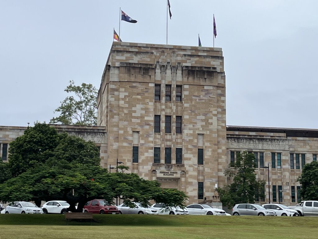 昆士兰大学福根史密斯楼砂岩建筑，澳大利亚国旗在晨风中飘扬。前景草地和树木，布里斯班阴天清晨的宁静校园氛围.