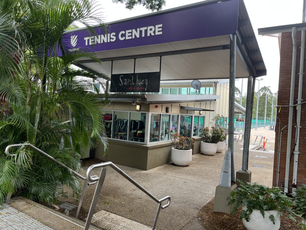 昆士兰大学网球中心正面入口，紫色背景配白色文字。五级台阶通向球场，右侧视角看到网球用品店和后方圣露西咖啡馆的户外桌椅。前景有棕榈树和树木，人行道上的小咖啡馆广告牌。