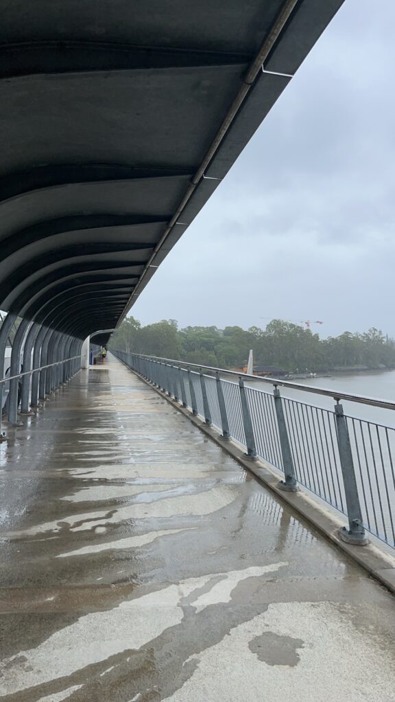 从绿桥步行道回望来路，轻雨中，灰色调的河景。左侧有弧形金属顶棚，右侧金属栏杆外可见布里斯班河。混凝土步道部分干燥，部分有雨水流过。远处桥身向消失点延伸。