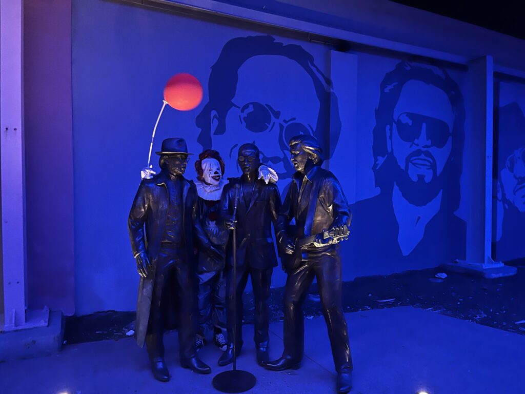 Bee Gees路夜间，蓝色灯光下，小丑潘尼怀斯手搭Bee Gees兄弟雕像肩膀，手持红气球