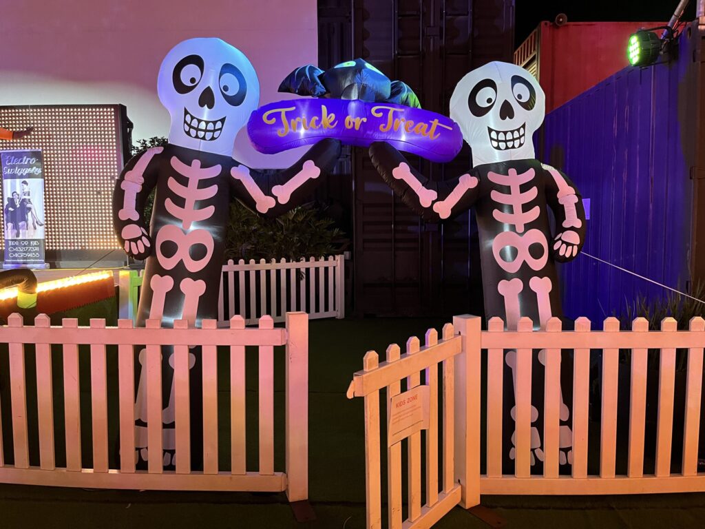 Eat Street夜市，白色木栅栏门开着，两侧高耸骷髅气球伸手撑起紫色「Trick or Treat」招牌，蝙蝠气球