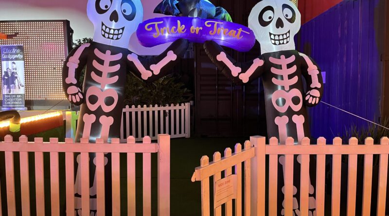 Eat Street夜市，白色木栅栏门开着，两侧高耸骷髅气球伸手撑起紫色「Trick or Treat」招牌，蝙蝠气球