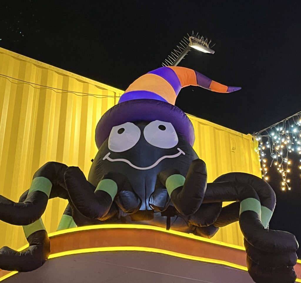 Eat Street夜市入口，黄色货柜屋顶上方，巨型充气黑蜘蛛戴紫橙巫师帽，绿色条纹腿，大笑眼