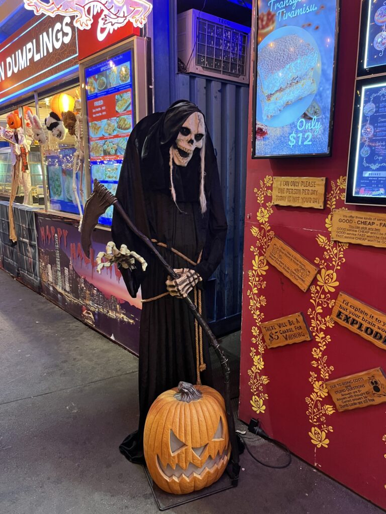 Eat Street夜市，黑色死神雕像手持大镰刀，脚边刻南瓜咧嘴笑，传统骷髅脸，身后甜品招牌提拉米苏$12