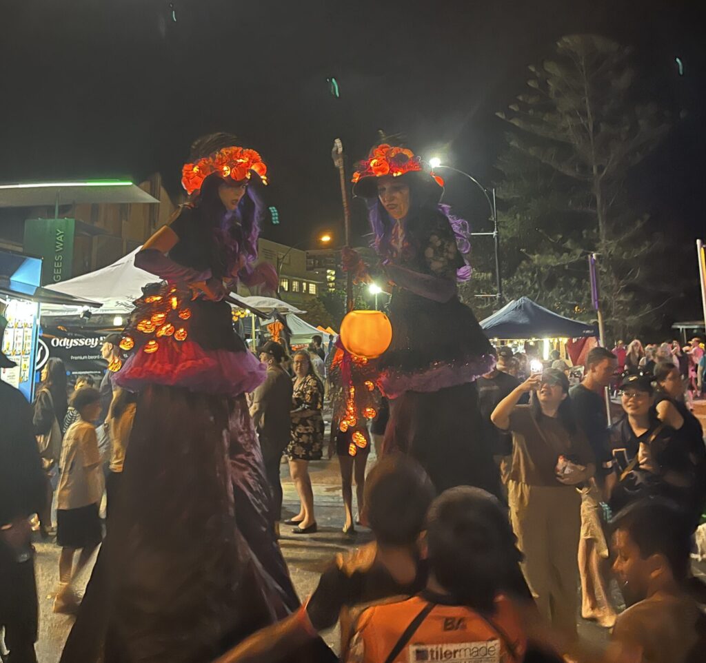 Redcliffe万圣节夜市，两位高跷女巫紫色黑色长裙，红发光帽，橙色南瓜魔法杖，给孩子发糖果