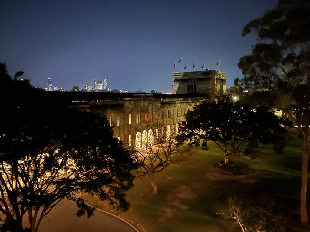昆士兰大学大庭院夜景，福根史密斯楼亮灯，草坪树木染黄光，远处CBD天际线