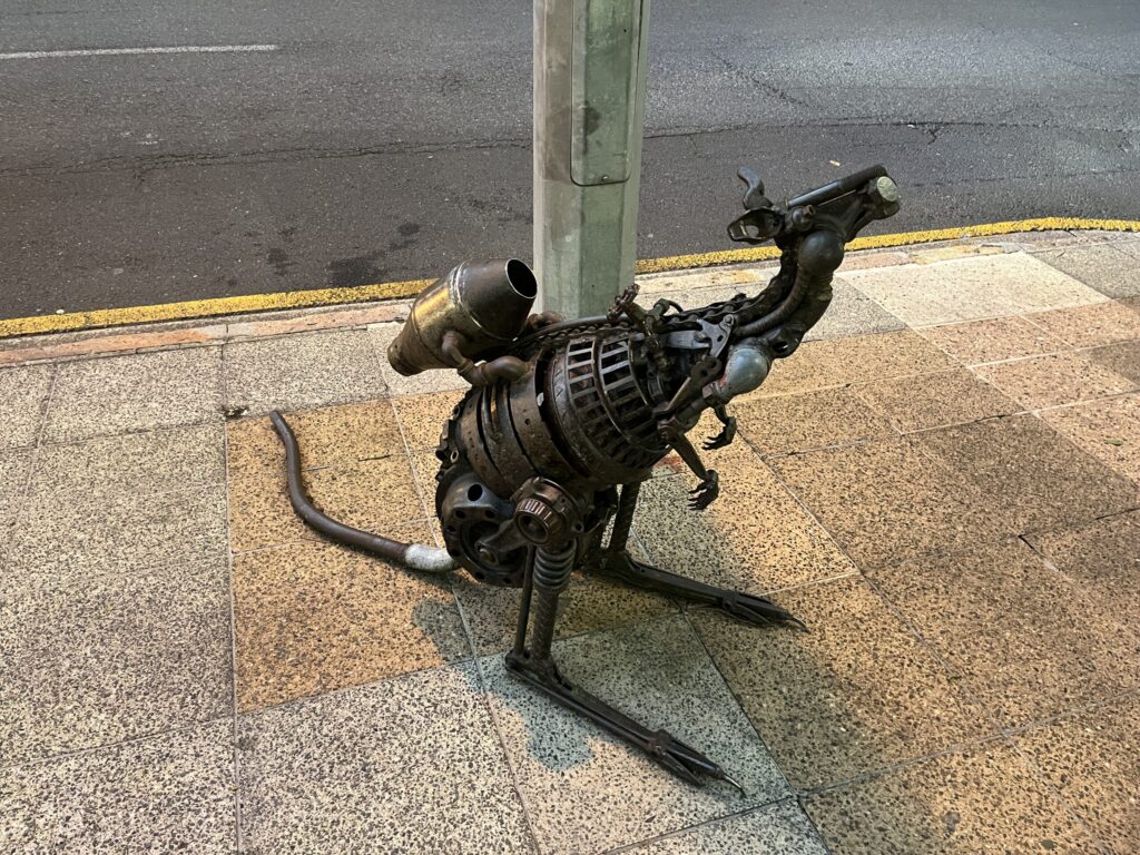 布里斯班乔治街金属袋鼠雕塑，站立于路边灯柱旁，头部倾斜好奇，背部有火箭喷气包细节