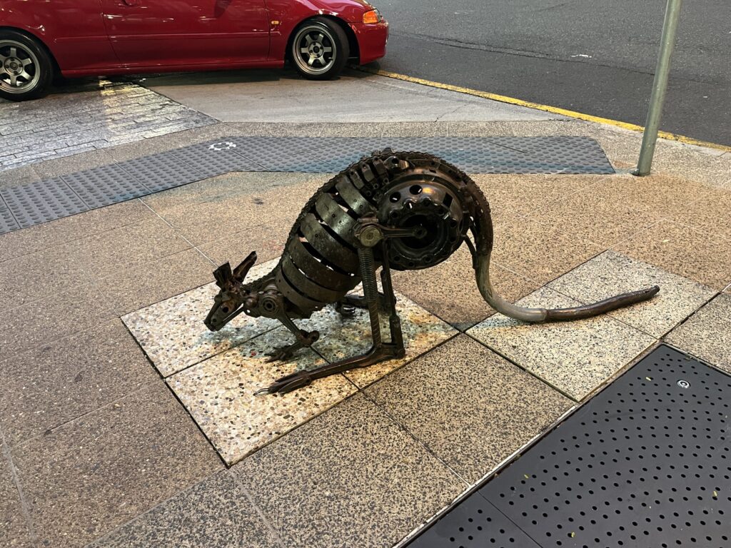 布里斯班乔治街金属袋鼠雕塑，觅食姿态位于人行道车道旁，红色汽车即将转弯驶出