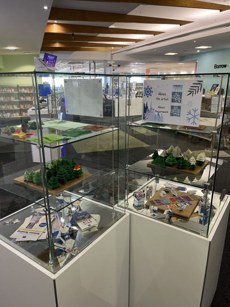 图书馆内两个方形玻璃展柜，白色底座，三层陈列：顶层折纸原料，中层森林景观，底层宣传模型纸鹤星星