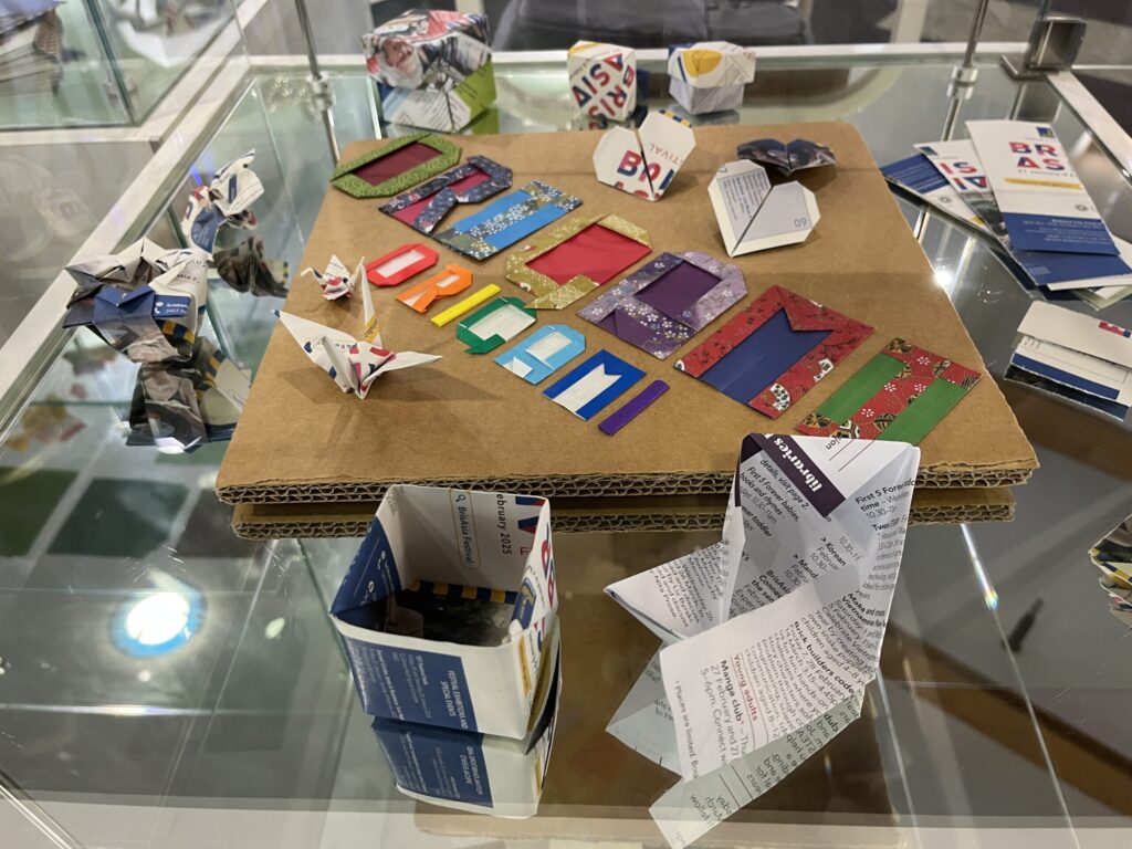 图书馆折纸展柜底层，棕色纸板中央，彩色折纸字母拼出ORIGAMI/origami，周围蓝白色宣传册折纸模型
