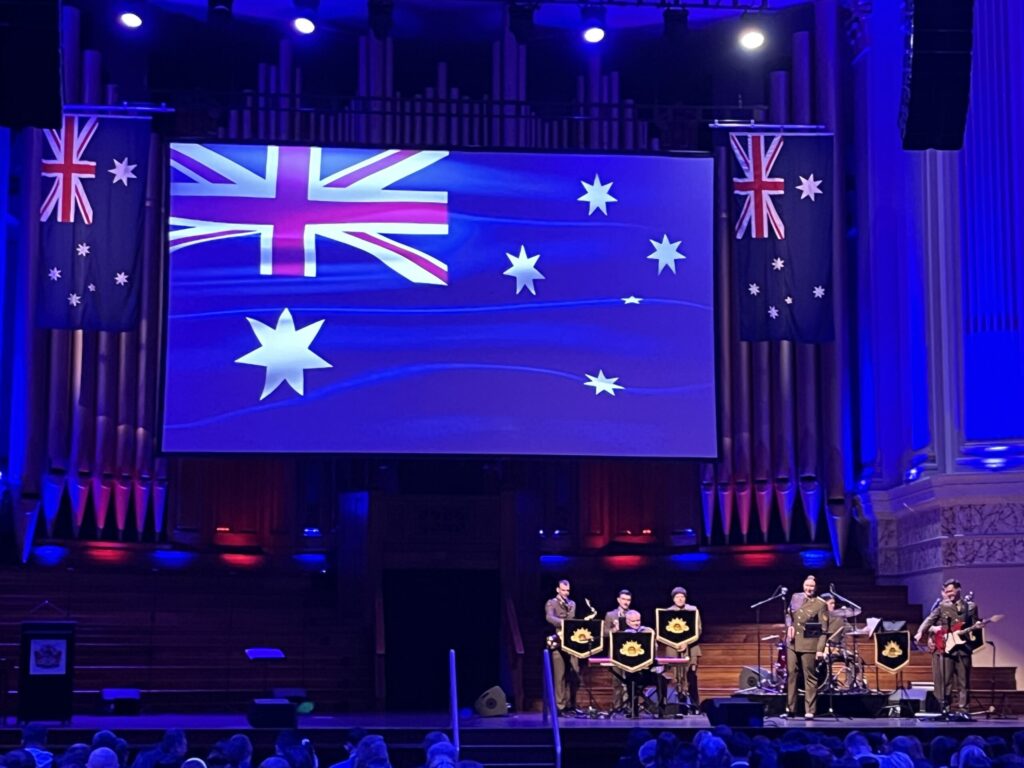 布里斯班市政厅礼堂舞台，背景大型管风琴（乐器），悬吊投影幕澳洲国旗，两侧实体国旗，左侧讲台，右侧乐队，前景观众头顶