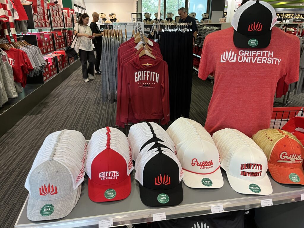 格里菲斯大学纪念品店，前台运动帽，左侧红色GU商品墙，红色卫衣，右侧红色T恤人台，后方货架毕业熊