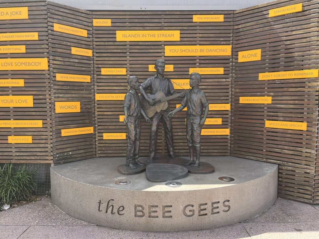 比吉斯之路少年青铜雕像，三人演奏，舞台刻乐队名，后方木格栅与金曲词牌