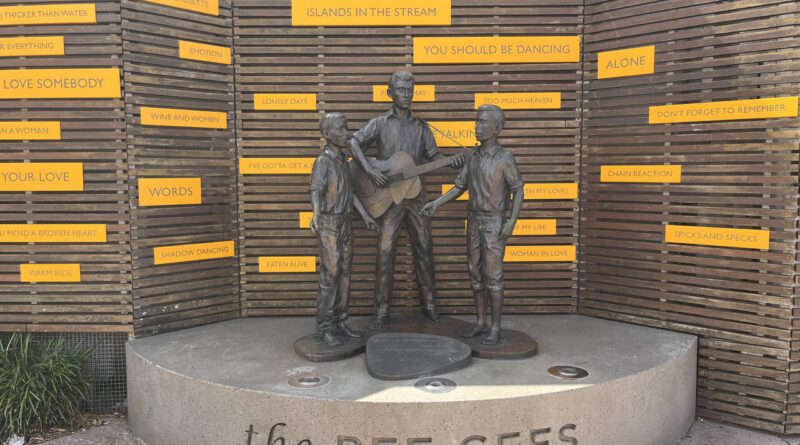 比吉斯之路少年青铜雕像，三人演奏，舞台刻乐队名，后方木格栅与金曲词牌