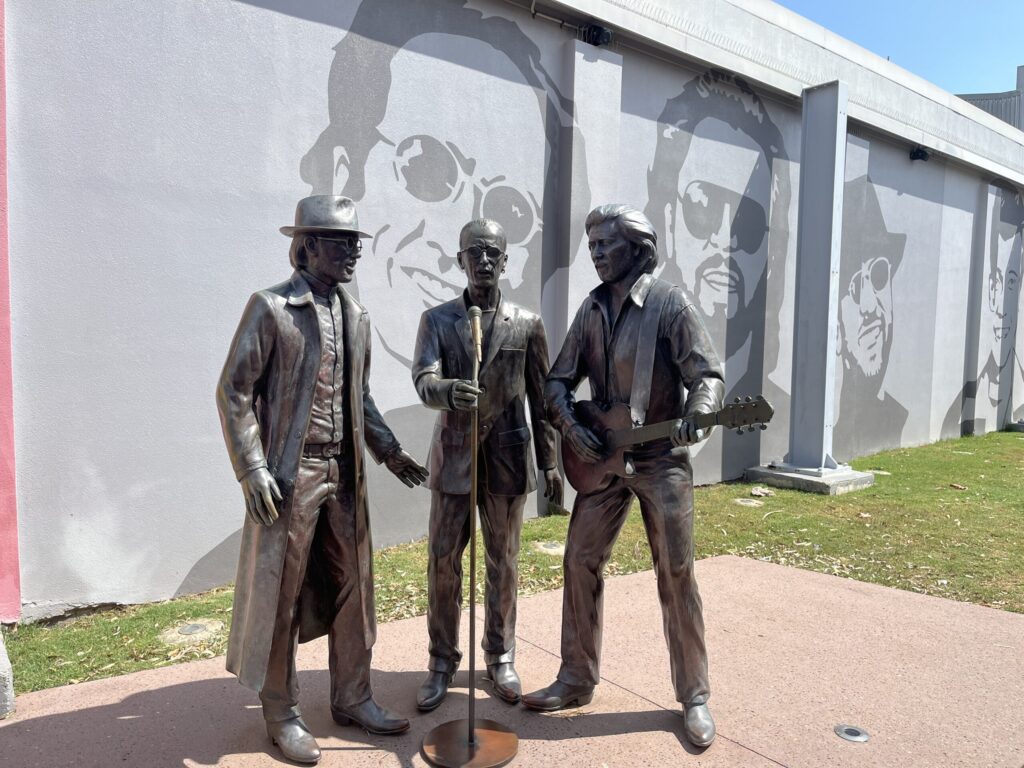 比吉斯之路真人大小的青铜雕像，一人弹吉他，一人握麦克风架，灰色墙面上有深灰色头像