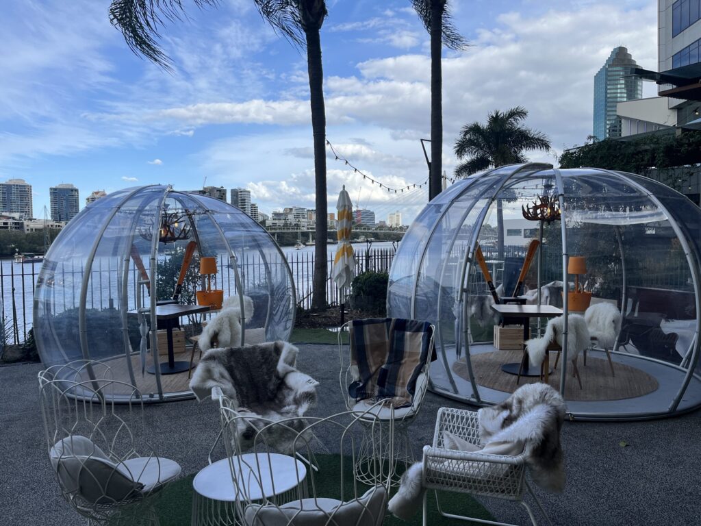 布里斯班海关大楼户外用餐区，透明塑料冰屋，内设餐桌羊皮椅，河景棕榈树蓝天白云