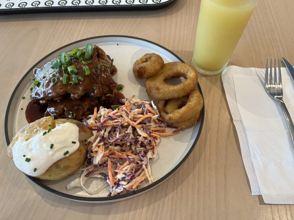 木桌上一盘烤猪肋排，配烤土豆、卷心菜沙拉、炸洋葱圈，菠萝汁，刀叉