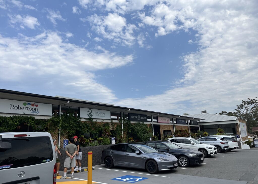 罗伯逊村购物中心全景，一排停车位，三人在木格栅入口前，店铺招牌，罗伯逊村标志，蓝天白云