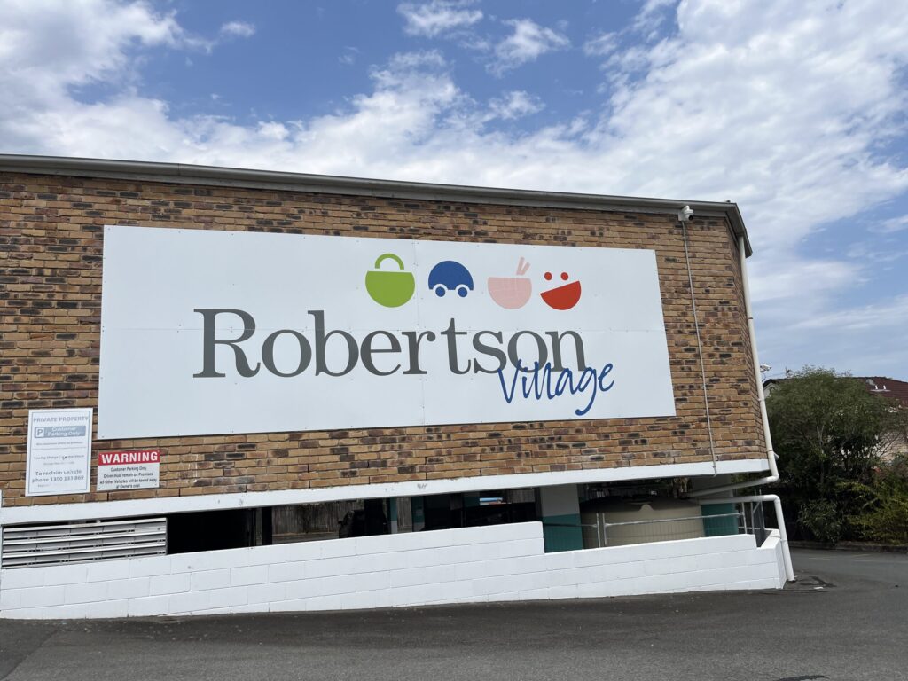 罗伯逊村建筑外墙招牌，棕色砖墙，白色「Robertson」罗马字体，蓝色手写体「village」，上方购物袋、汽车、面碗图标