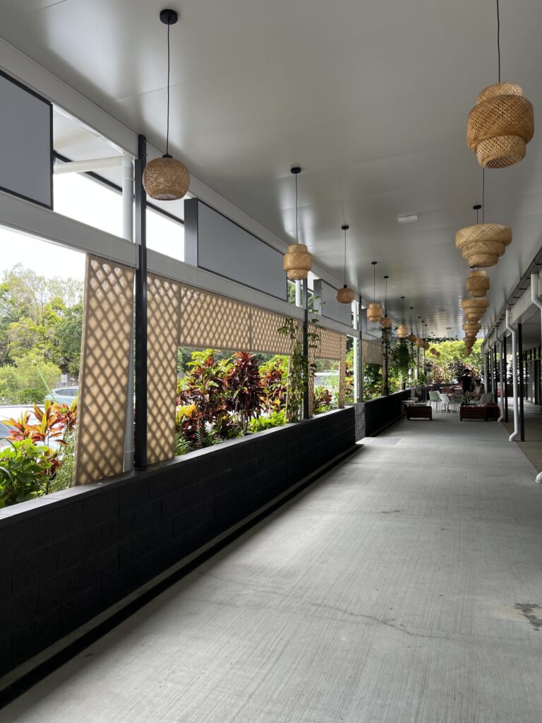 罗伯逊村户外用餐区，左侧木格栅，右侧店铺，远处沙发咖啡椅，两排稻草灯罩吊灯