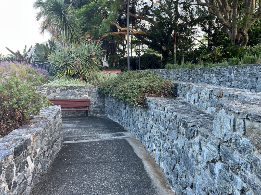 库萨山植物园U形弯道缓坡，棕色木椅在坡底，右侧石墙高台石凳，上方另一木椅