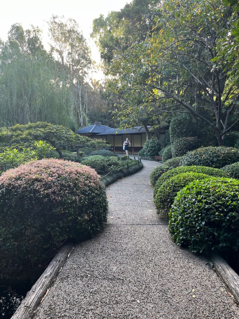 日本花园池塘边小径，圆形修剪灌木，女子走向镜头，遮阳长椅
