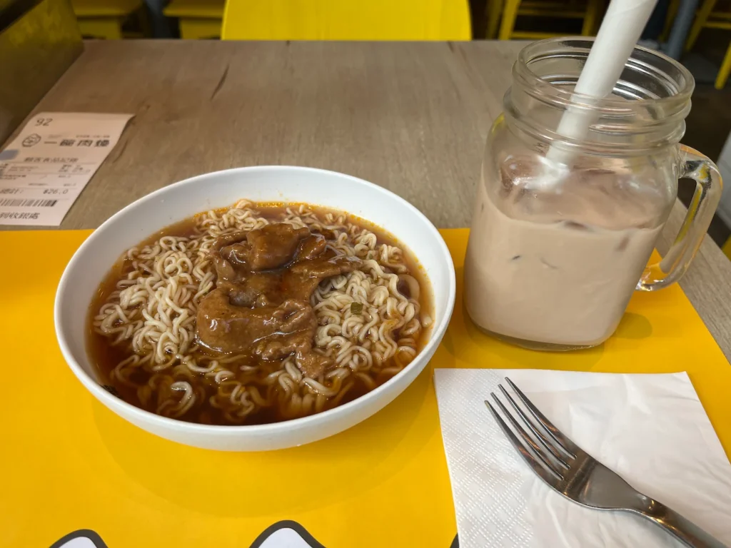 咖啡桌中心一碗沙爹牛肉方便面，左侧冰奶茶，餐巾与叉子在前