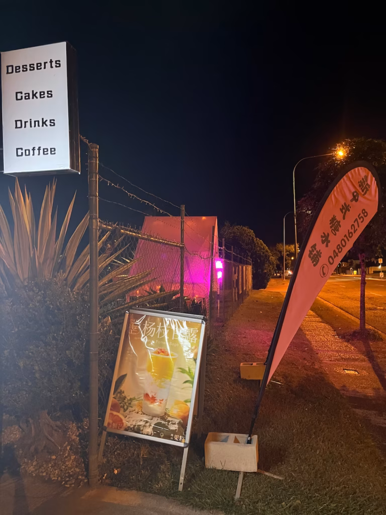 Pinelands Road夜晚，黄色街灯，铁丝网上方白黑招牌「desserts cakes drinks coffee」，草地A字牌芒果西米露图，粉色旗帜牌，远处小屋粉色招牌发光