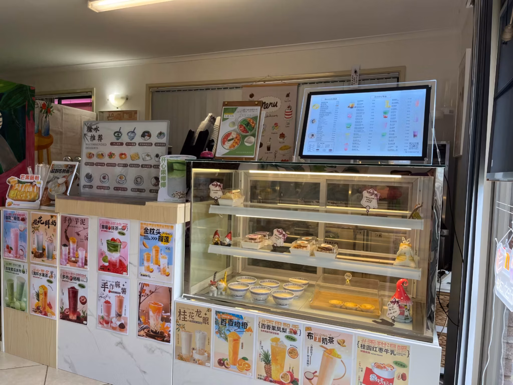 Landy's Sweet House进门左侧，主柜台与蛋糕展示柜，彩色饮品海报覆盖柜台正面，灯光菜单屏，后方饮品工作台
