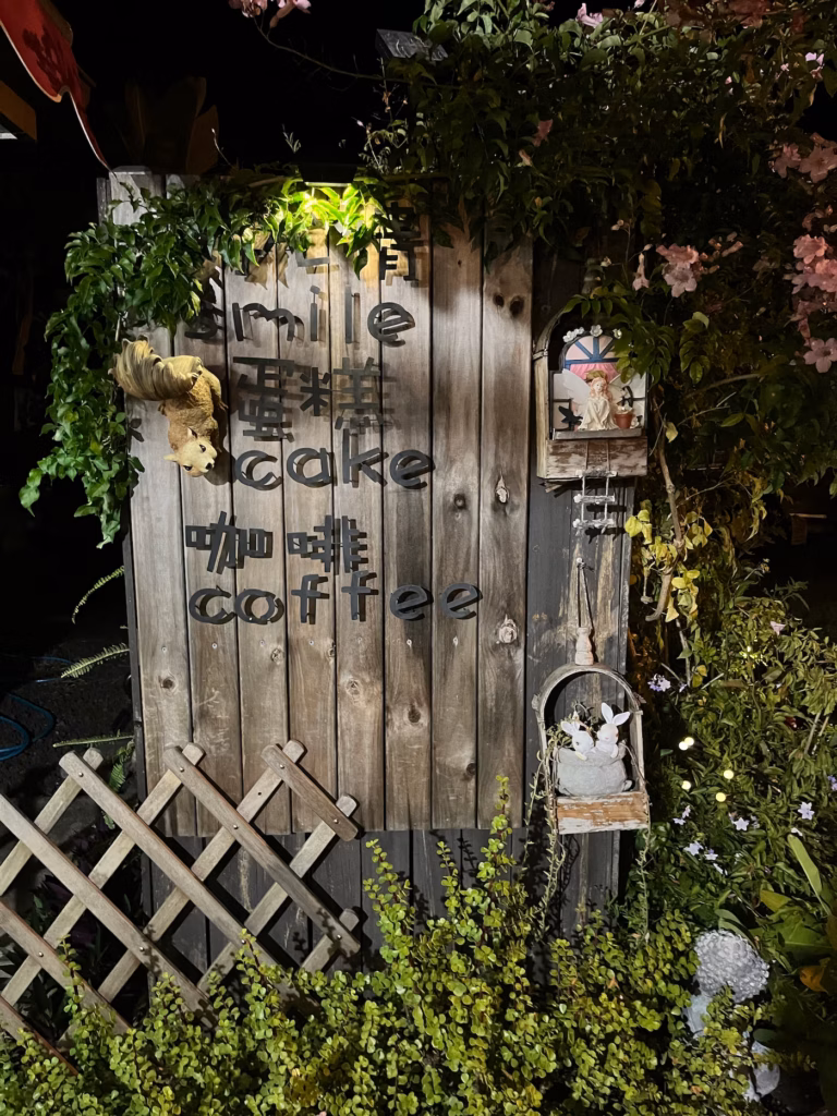 Landy's Sweet House木栅栏，黑字「Smile, Cake, Coffee」，花丛松鼠、小屋、兔、石像，白紫花盛开