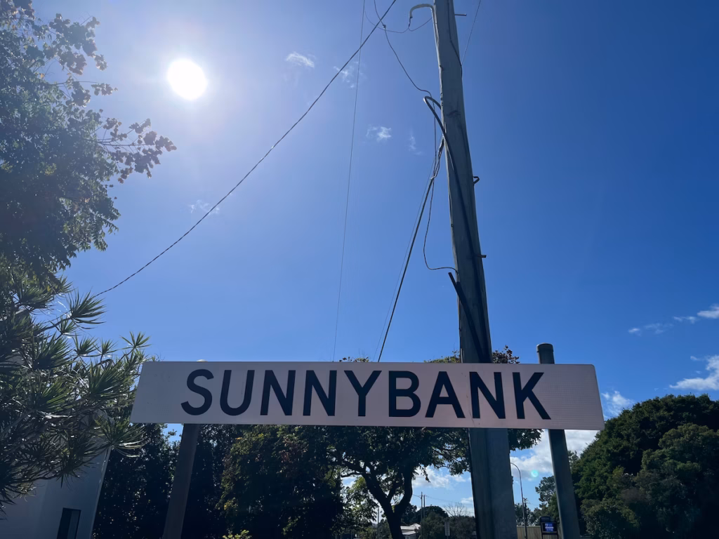 Sunnybank郊区路标，白色黑字，近景，下方绿树冠，右侧木电杆，左后方正午太阳射线照亮标志