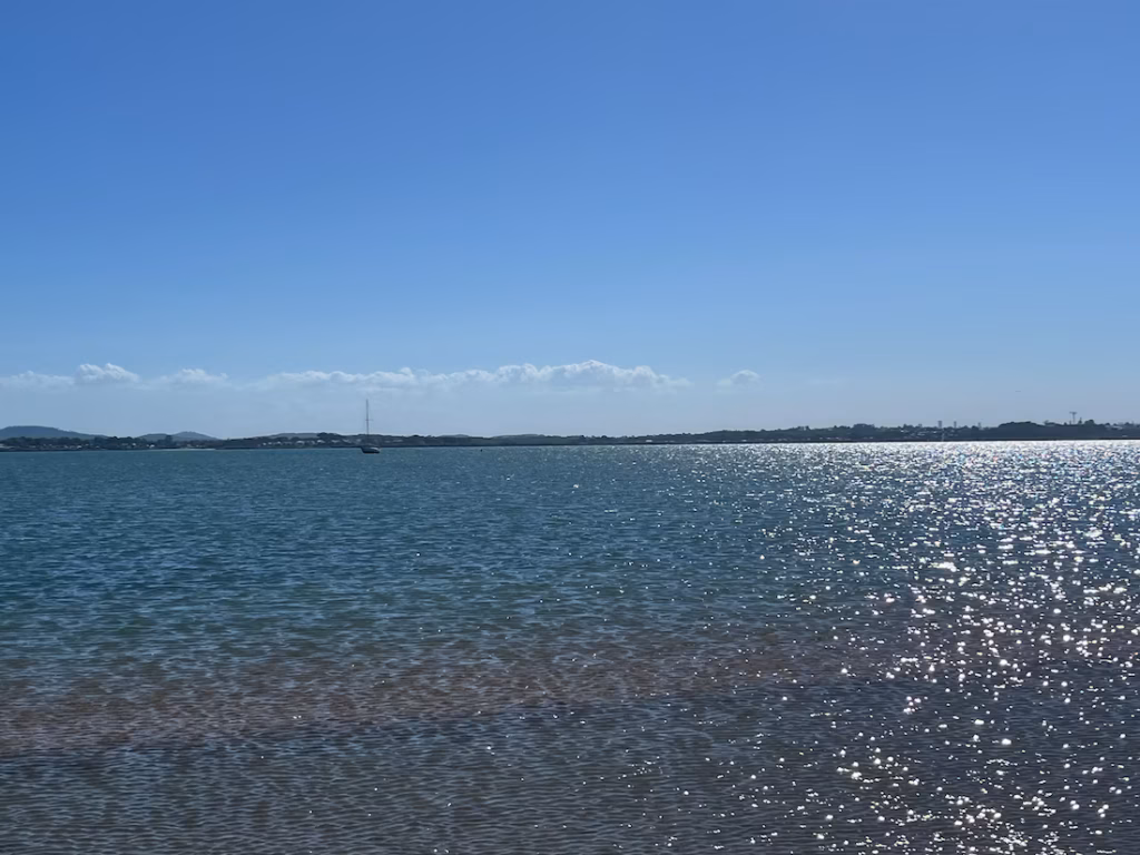 威灵顿角沙洲浅浪外，帆船在远方水面，涟漪微光，棕色沙底清晰可见，远处陆地白云，蓝天