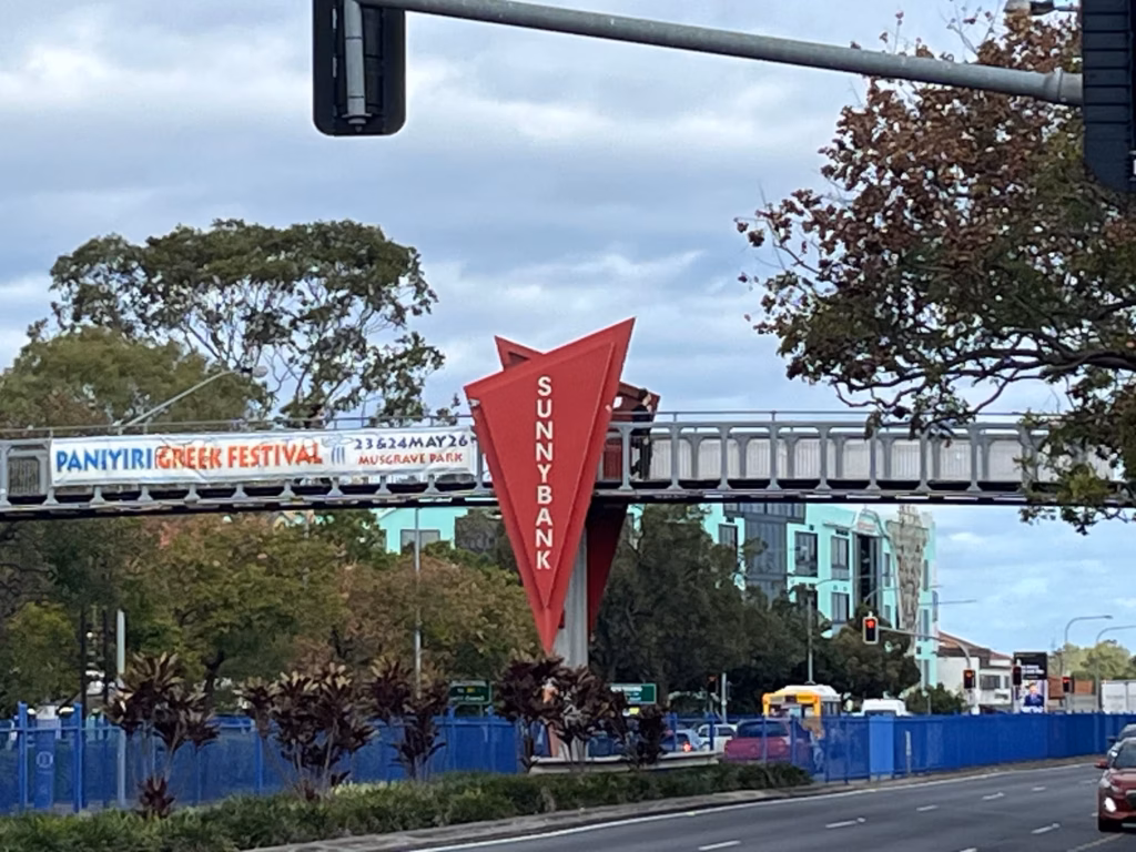 Sunnybank Plaza侧，红色Sunnybank天桥标志，前景Mains Road，中央隔离带蓝色护栏，红绿灯，右侧树冠，背景Market Square建筑