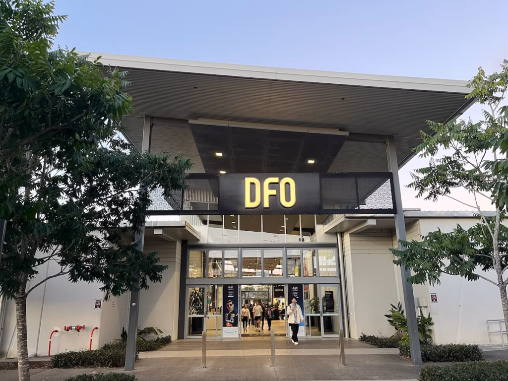 布里斯班DFO入口，灰白建筑，大玻璃门，两侧树木，拱形黑色金属框架，白色霓虹「DFO」大字，蓝灰色天空