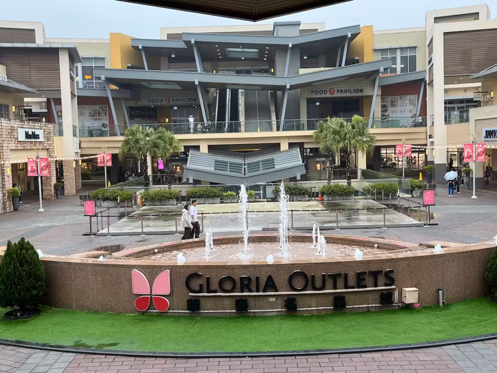 台北Gloria Outlets开放式庭院，喷泉带中心名称与蝴蝶标志，背景美食广场餐厅，阴天工作日
