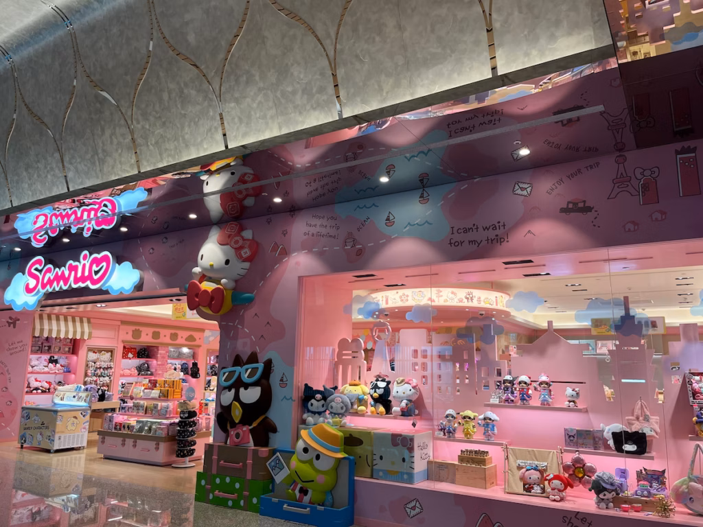 台北机场Hello Kitty商店，粉红外墙，大型橱窗展示玩具公仔，入口上方Kitty乘飞机，红蝴蝶结作螺旋桨，地上两同伴拎行李箱挥手