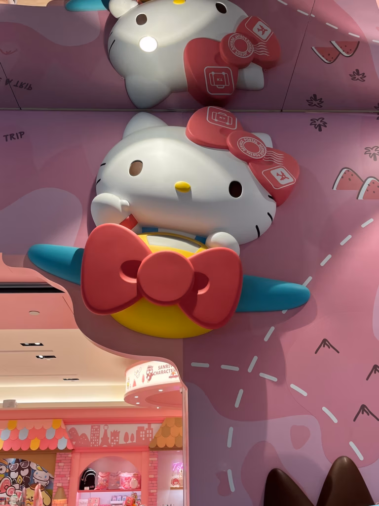 台北机场Hello Kitty商店入口上方，黄色飞机蓝色机翼，粉红蝴蝶结螺旋桨，Kitty戴蝴蝶结护照印章，挥手