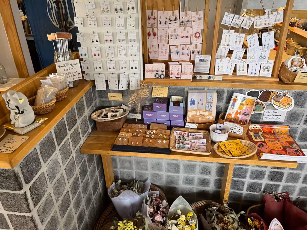 Runcorn 咖啡馆室内，长台右侧展示本地手作首饰与工艺品，台下干花束，左角灰色招财猫